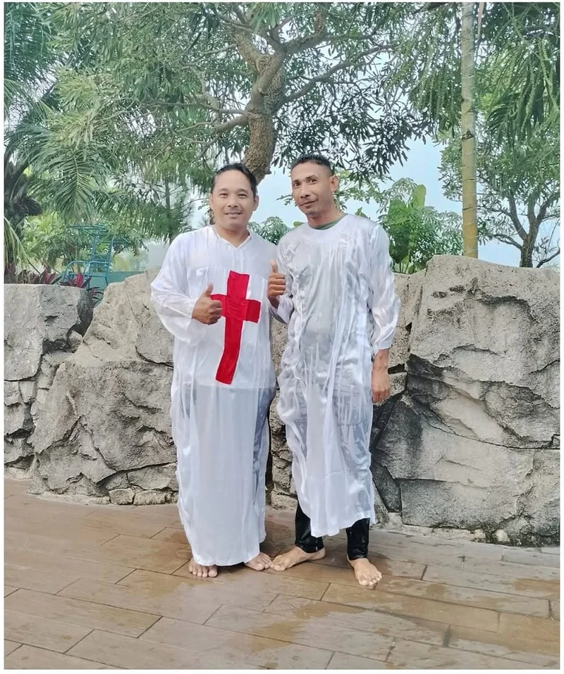 Dua orang mengenakan pakaian baptisan putih berdiri di luar ruangan dengan latar batu dan dedaunan.