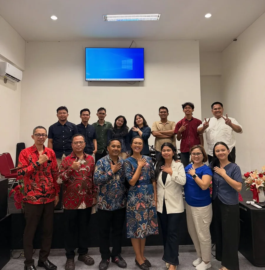 Kelompok pelayanan GBI SCM berfoto bersama di sebuah ruangan kecil dengan layar di dinding belakang.